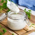 45 degree angle shot of small glass jar on a wooden cutting board filled with Greek yogurt ranch dressing. A spoon is hovering above filled with the dressing.
