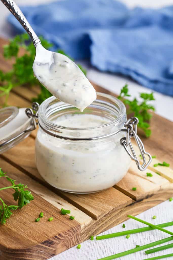 45 degree angle shot of small glass jar on a wooden cutting board filled with Greek yogurt ranch dressing. A spoon is hovering above filled with the dressing.