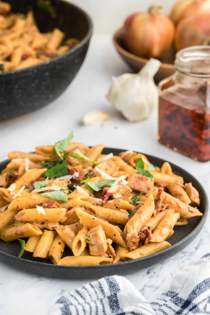 A dark gray plate with Marry Me Chicken Pasta in it garnished with fresh basil, and shredded parmesan cheese. In the background there is a bottle of sun-dried tomatoes, a garlic bulb, a wooden bowl with onions and garlic, and a frypan with Pasta in it waiting to be served