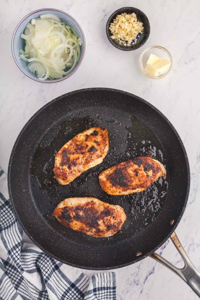 Over head shot of chicken breasts searing in a frypan. Top left corner is a bowl of sliced onions, and top right corner is a pinch bowl of minced garlic and a pinch bowl of butter, next ingredients to be added in the recipe.