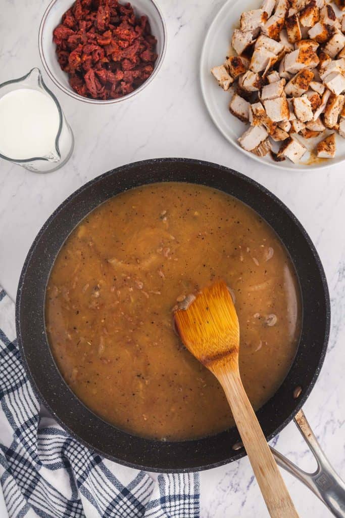 Overhead shot of frypan being deglazed with chicken stock. Next ingredients to be added are above, upper right corner a plate of the cooled and chopped chicken, upper left corner contains a cup of cream and a bowl of chopped sundried tomatoes