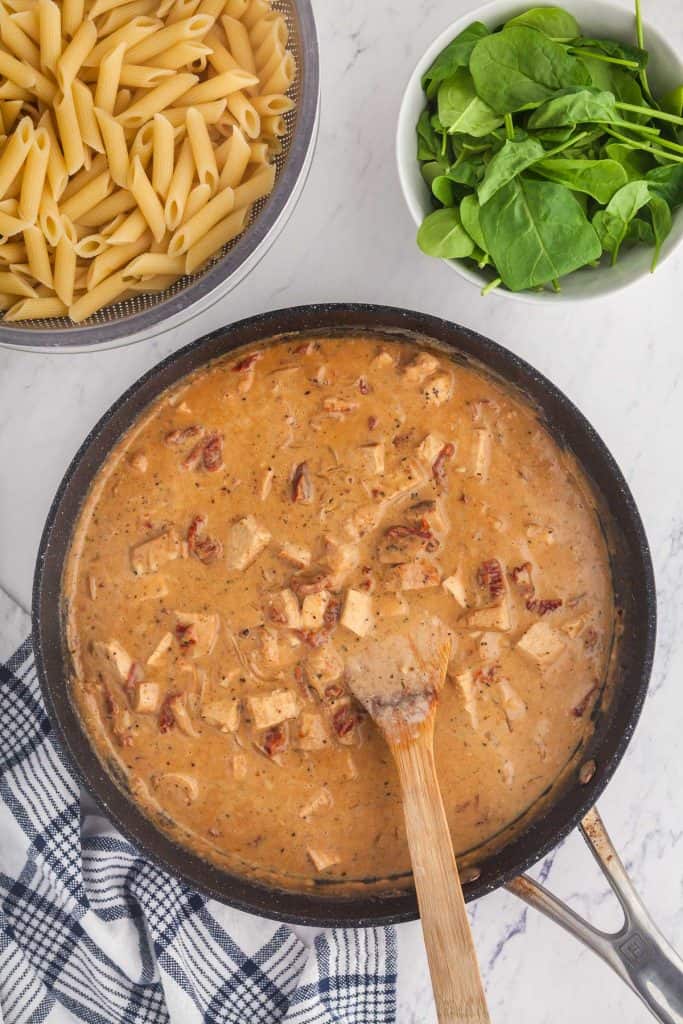 Overhead shot of the frypan containing the sauce for Marry Me Chicken Pasta. In the upper left corner is a big bowl of penne pasta prepared al dente and in the right upper corner is a bowl of baby spinach both ready to be added in the next step.