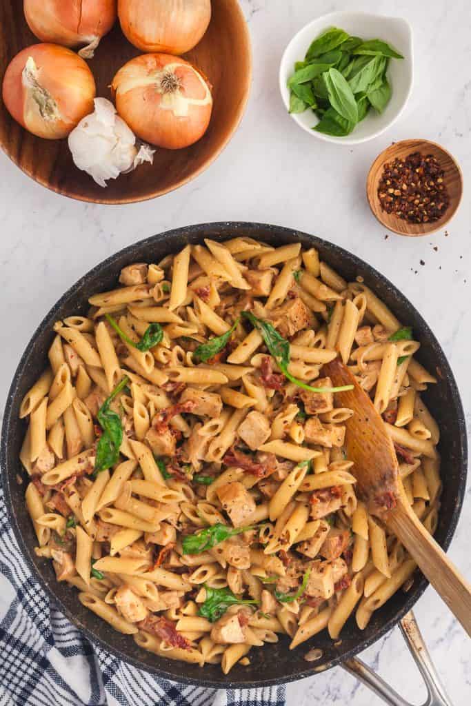 Overhead shot of Marry Me Pasta Chicken finished in a large frypan. Upper left corner is a wooden bowl with onion and garlic, and the right hand corner has two pinch bowls, one with basil leaves and the other with crushed chili flakes.