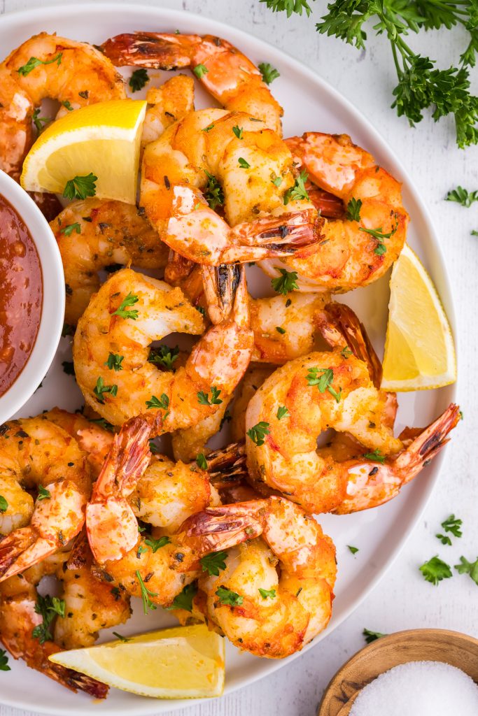 Close up overhead shot of a white plate filled with air fried pink shrimp coated in orange colored seasoning served alongside a red shrimp sauce and some fresh lemon wedges.