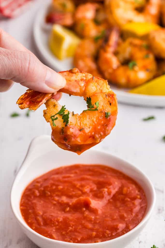 Close up side shot of a air fryer cooked shrimp being dipped in shrimp cocktail sauce. Background is of a big plate loaded with more shrimp.
