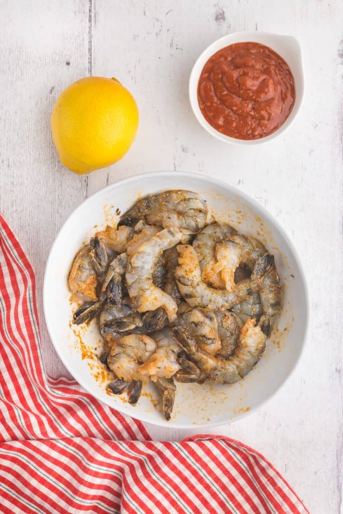 Overhead shot of raw shrimp marinating in bowl with marinade. Top left corner has a lemon and top right corner has red cocktail sauce.