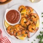 Overhead shot of Air Fryer Frozen shrimp nicely cooked and curled up on a white serving plate with a small bowl of red seafood dipping sauce and lemon wedges scattered around the plate. Above the plate to the right is a small bowl of lemon wedges, and to the left is a wooden bowl with fresh garlic and onion.