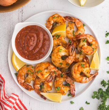 Overhead shot of Air Fryer Frozen shrimp nicely cooked and curled up on a white serving plate with a small bowl of red seafood dipping sauce and lemon wedges scattered around the plate. Above the plate to the right is a small bowl of lemon wedges, and to the left is a wooden bowl with fresh garlic and onion.