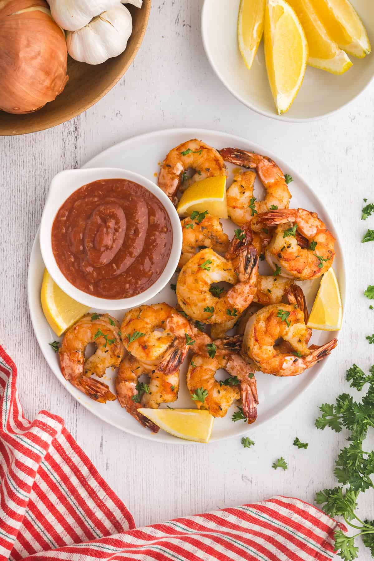 Overhead shot of Air Fryer Frozen shrimp nicely cooked and curled up on a white serving plate with a small bowl of red seafood dipping sauce and lemon wedges scattered around the plate. Above the plate to the right is a small bowl of lemon wedges, and to the left is a wooden bowl with fresh garlic and onion.