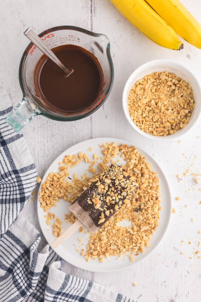 overhead shot showing a chocolate dipped banana popsicle laying in a plate of crushed peanuts so that it becomes coated with peanuts.