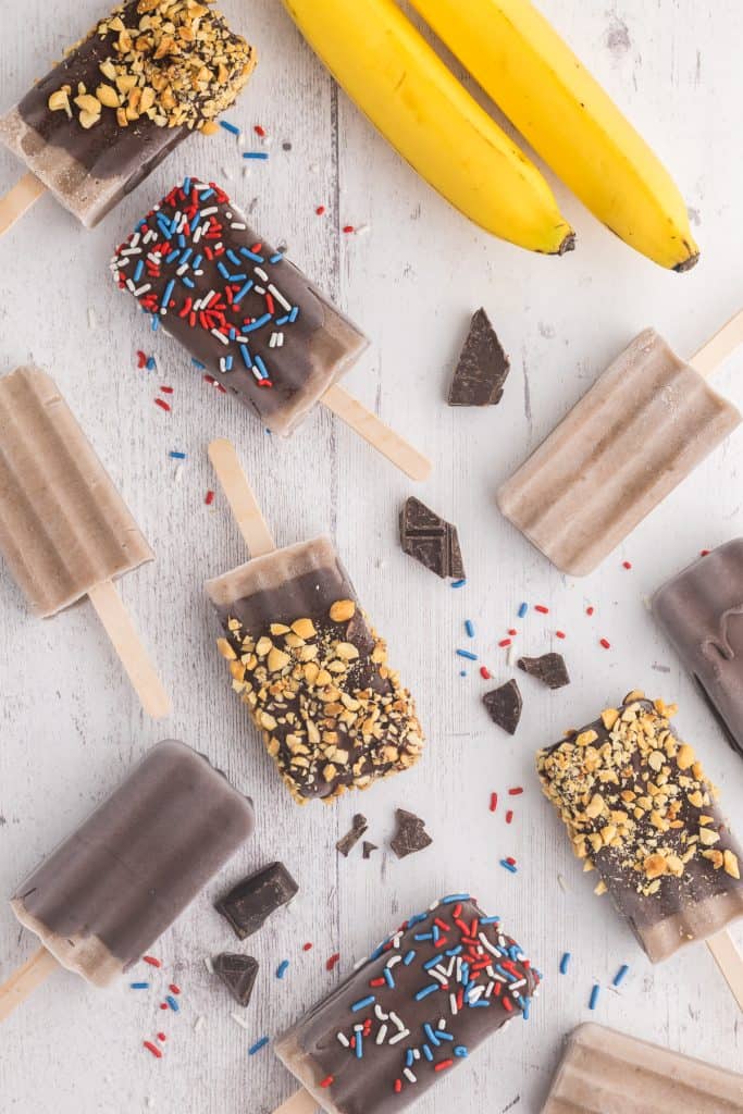 overhead photo of variously decorated banana popsicles laying on a white wooden table. Some are plain, some dipped in chocolate, some dipped in chocolate and rolled in crushed peanuts, and some dipped in chocolate and rolled in red white and blue sprinkles