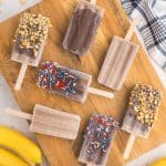seven banana popsicles laying askew on a wooden cutting board. Some are dipped in chocolate with crushed peanuts, others dipped in chocolate with red white and blue sprinkles and few are plain.