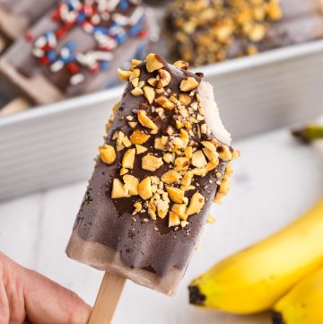 Banana popsicle being held up after it has been dipped in chocolate, rolled in crushed peanuts, and a bite taken out of it.