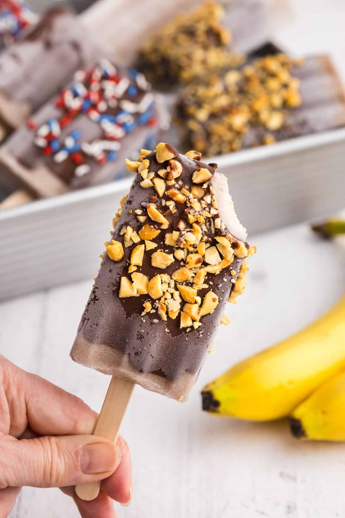 Banana popsicle being held up after it has been dipped in chocolate, rolled in crushed peanuts, and a bite taken out of it.