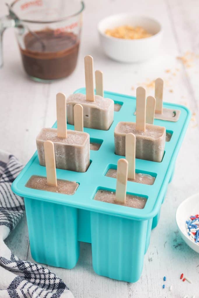 side shot looking at a blue silicone popsicle mold with frozen banana popsicles inside. A few of the popsicles and partially pulled out. In the background is a pyrex measuring cup filled with magic shell and a white bowl filled with crushed peanuts
