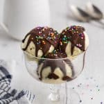 Glass dessert dish filled with three scoops of vanilla gelato covered in chocolate magic shell with multicolored sprinkles on top. In the background is a small jug of liquid magic shell and two spoons.