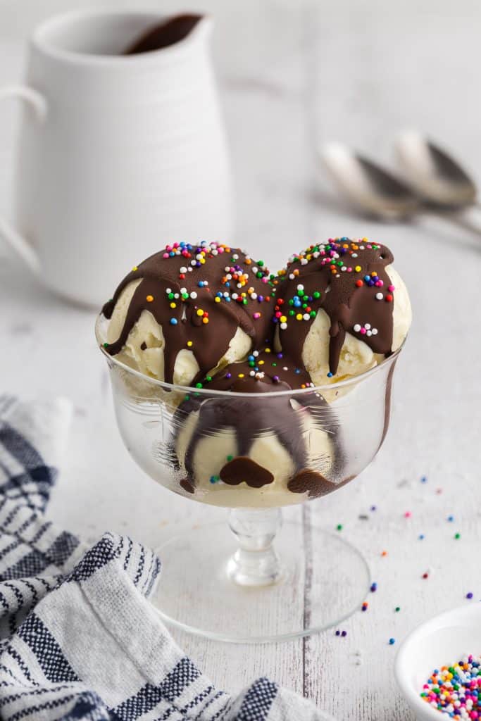 Glass dessert dish filled with three scoops of vanilla gelato covered in chocolate magic shell with multicolored sprinkles on top. In the background is a small jug of liquid magic shell and two spoons.
