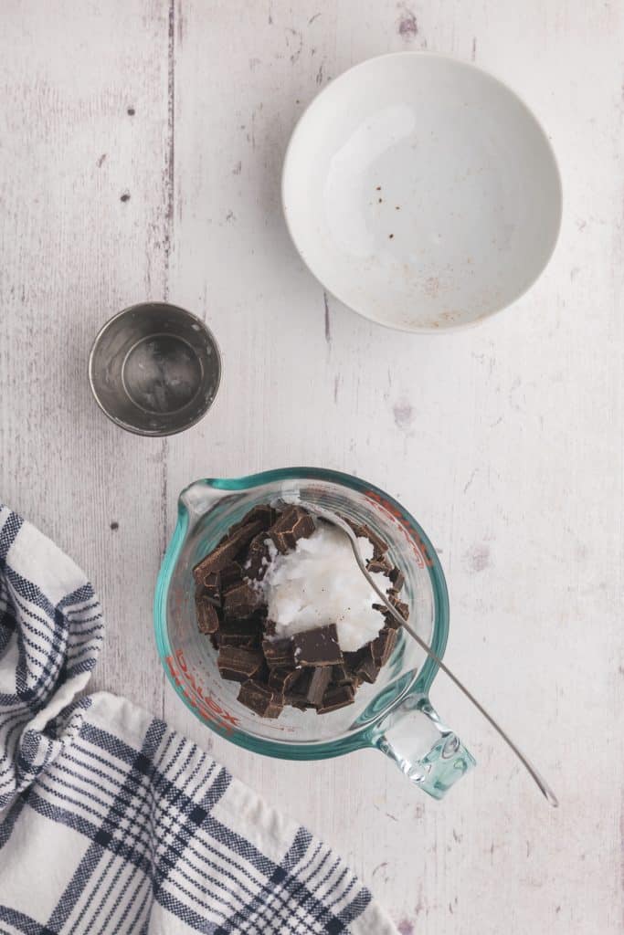 Overhead shot of a pyrex measuring cup with chopped chocolate and coconut oil inside. A spoon is in the measuring cup, and the empty pinch bowl of coconut oil, and empty bowl of chocolate sit above the measuring cup.