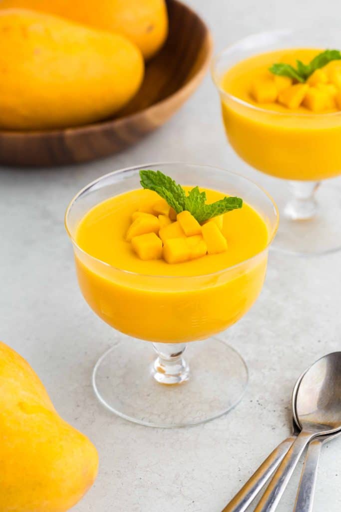 Glass dessert bowl filled with a bright yellow mango pudding garnished with diced mangoes and a small sprig of mint.