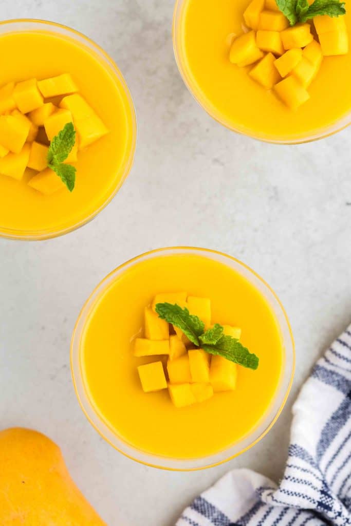 Overhead shot of mango puddings which have been garnished with diced mango a sprig of fresh mint. Three pudding cups in total, one in the middle, one in the upper left corner and one towards the upper right corner.