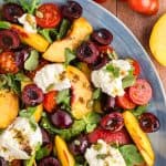 Peach burrata salad on a large platter taking up the whole photo except the top right corner which has cherry tomatoes and a slice peach peeking in.