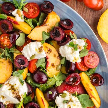 Peach burrata salad on a large platter taking up the whole photo except the top right corner which has cherry tomatoes and a slice peach peeking in.