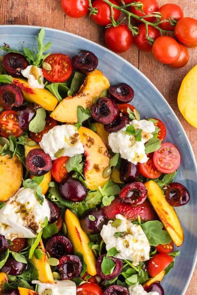 Peach burrata salad on a large platter taking up the whole photo except the top right corner which has cherry tomatoes and a slice peach peeking in.
