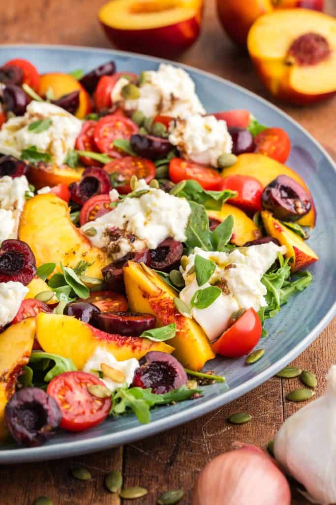 Peach burrata salad bursting with fresh peaches, cherries, cherry tomatoes, burrata cheese and fresh basil and oregano. A light drizzle of a balsamic vinaigrette dressing. The top right corner has a two halves of a sliced peach peeking in, the bottom right corner has a garlic bulb and shallot peeking in.