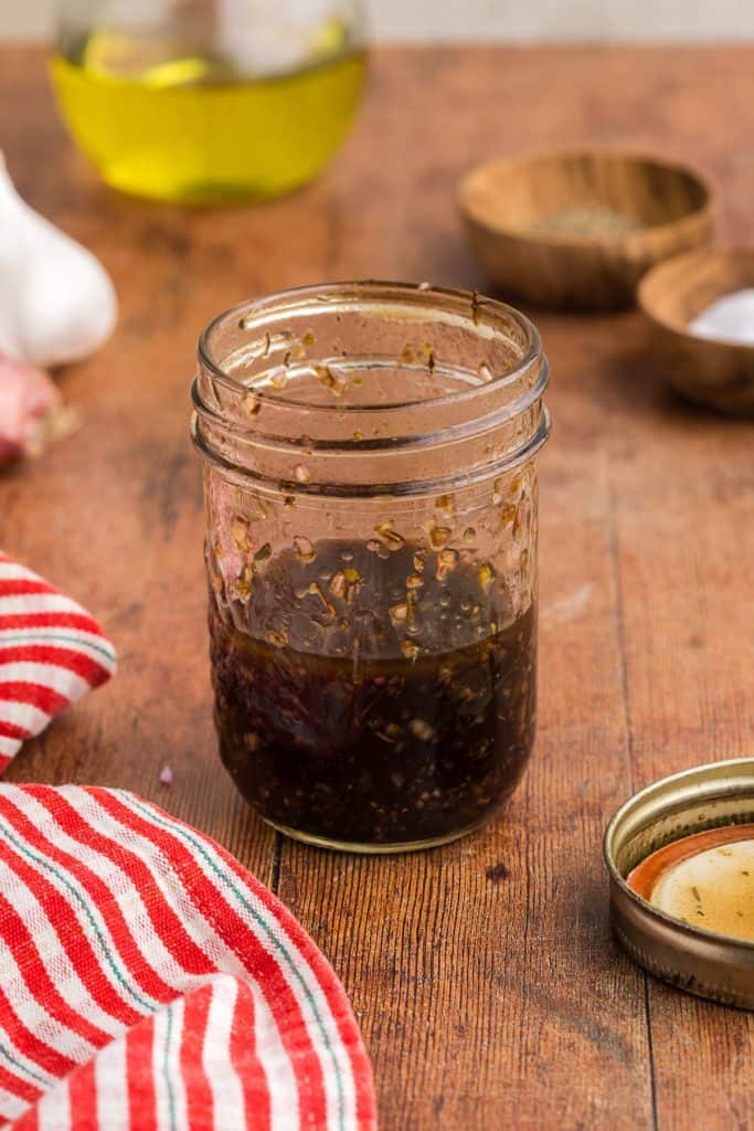 Side shot of a small jar filled with the ingredients for the vinaigrette. Jar has been shaken and the lid is off and sitting to the right. A bulb of garlic and a shallot are peeking in in the top left corner, and wooden pinch bowls of salt and pepper are in the top right hand corner