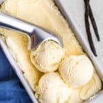 overhead view of three scoops of ice cream sitting a top of a pint of vanilla gelato in a metal loaf tin.  Two vanilla beans lay across each other to the right and a blue linen lays on the left of the ice cream tin.