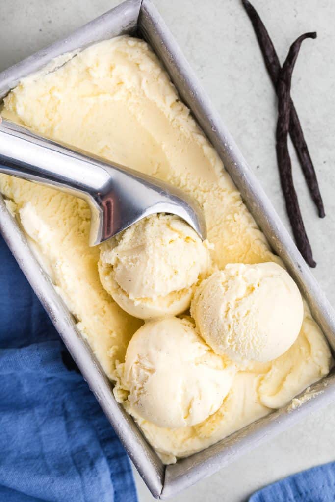Overhead shot of a metal loaf pan filled with creamy vanilla gelato.  An ice cream scoop has just made three balls of ice cream in the container.  A couple of vanilla beans are poking in to the scene in the upper right corner.