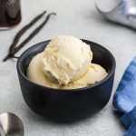Three scoops of vanilla gelato in a small black ceramic bowl. Vanilla beans on the side.