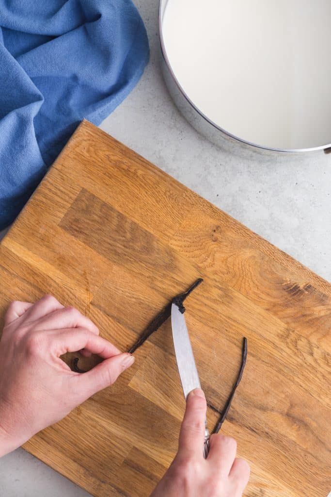 Overhead shot of a vanilla bean pod on a wooden cutting board having it's seeds scraped out with the dull side of a knife.