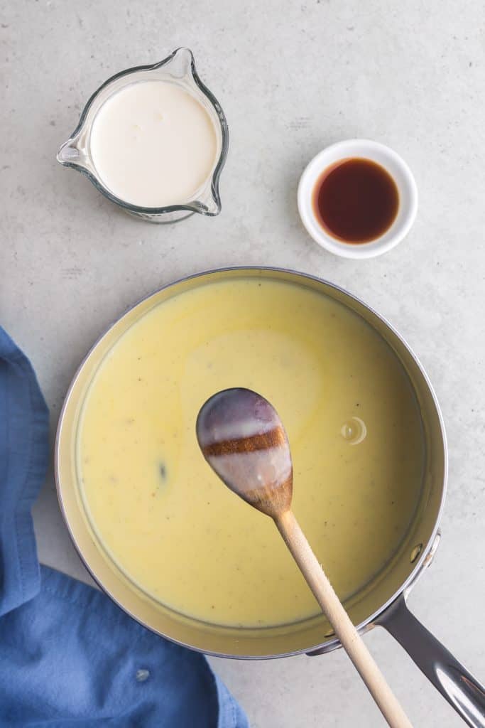 Overhead shot of the back of a wooden spoon showing the thickness of the custard - just enough to coat the back of the spoon. Below the spoon is the saucepan with the rest of the vanilla gelato custard.