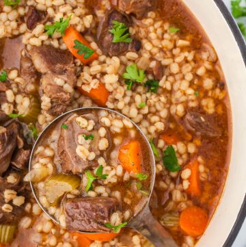 Very close up shot of Beef and Barley soup with a ladle lifting out a serving filled with a red beefy broth and big chunks of beef, carrots, and celery an dotted with lots of barley.