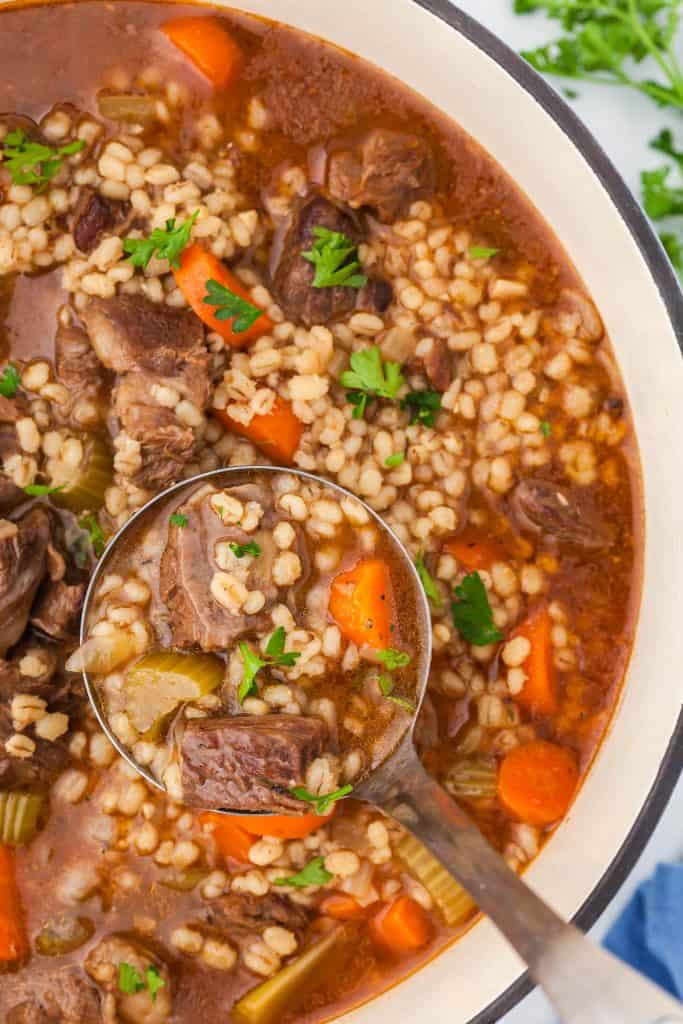 Very close up shot of Beef and Barley soup with a ladle lifting out a serving filled with a red beefy broth and big chunks of beef, carrots, and celery an dotted with lots of barley.