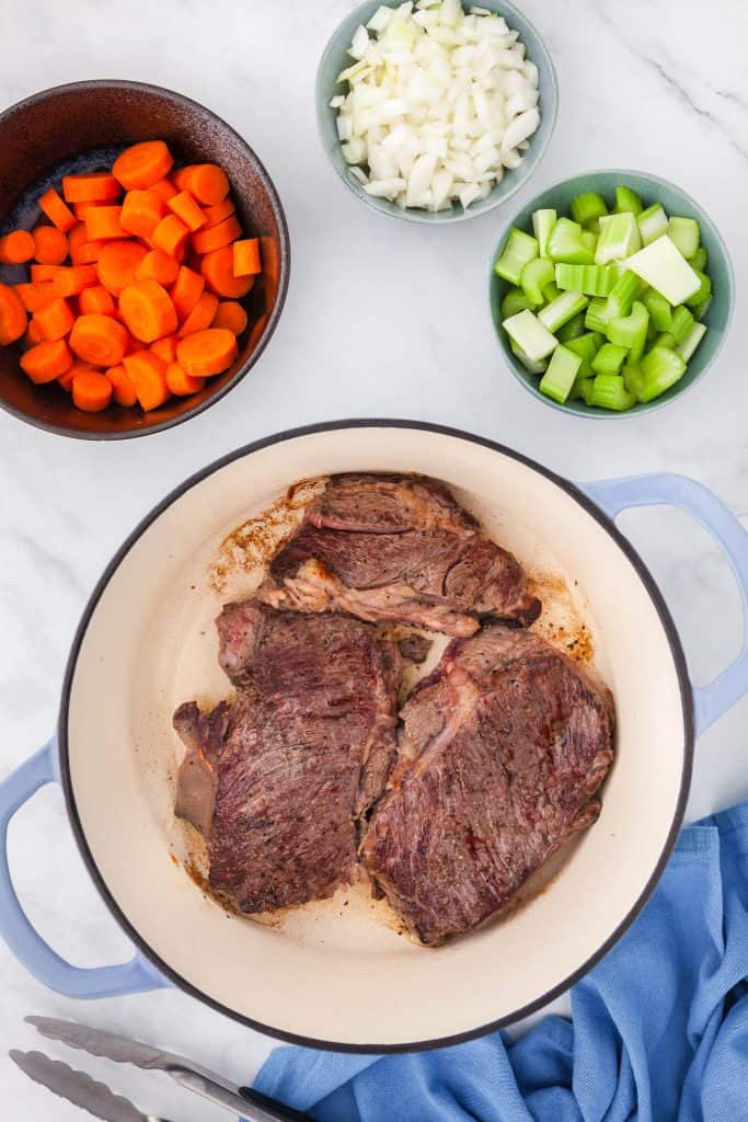 Overhead shot of beef steaks being seared in a Dutch oven. A bowl of carrots wait in the upper left corner, while bowls of onions and celery wait to be added in the upper right corner.
