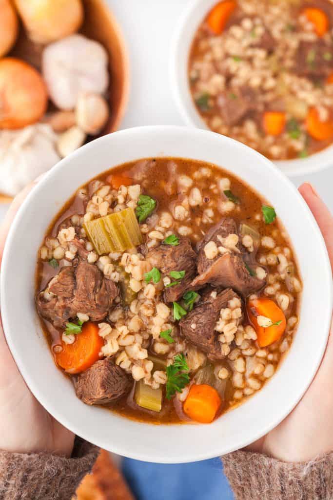 Close up of a bowl of chunky beef and barley soup being held between two hands. The background below has another bowl of beef barley soup peeking in from the top right and the top left is a wooden bowl filled with onions and garlic