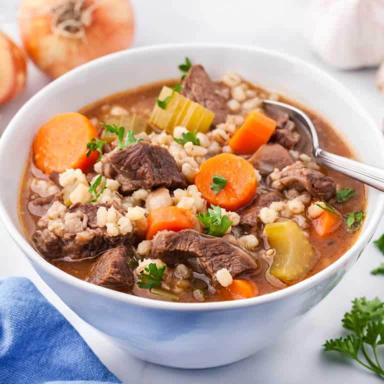 Close up side shot of a white porcelain bowl filled with beef and barley soup. There are big chunks of beef, carrots, and celery, with lots of barley, a sprinkling of fresh parsley, all in a rich beefy broth.