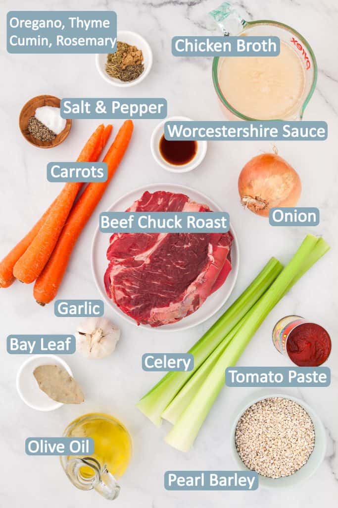 Overhead shot of all the ingredients necessary to make Beef Barley Soup. Each item is labelled with a blue label and white font. Ingredients needed are: Beef chuck roast, carrots, celery, onion, chicken stock, garlic, bay leaf, thyme, oregano, rosemary, cumin, tomato paste, pearl barley, Worcestershire sauce, salt and pepper