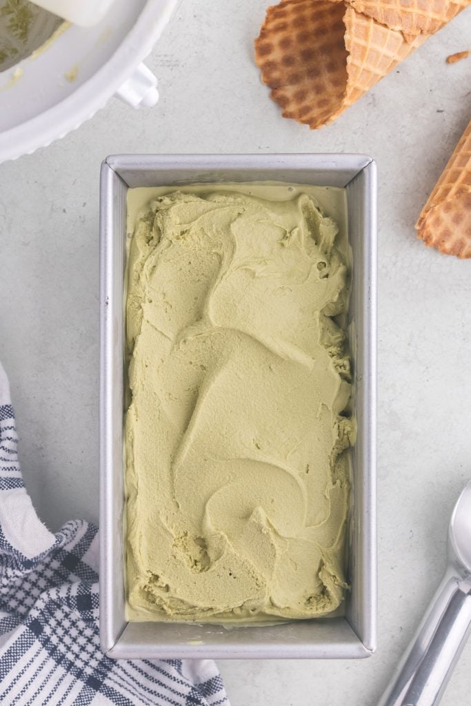 Overhead shot of green matcha ice cream in a silver metal loaf tin. The surface of the ice cream is swirly and ready to go in the freezer to firm up. Some waffle cones are scattered around the upper right hand corner, and an ice cream scoop is peeking in the bottom right hand corner