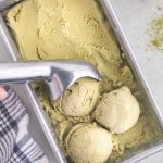 Overhead view of matcha ice cream in a metal loaf pan. Three scoops of ice cream have been pulled with the metal ice cream scooper and sit near the bottom of the container. There is a sprinkling of matcha powder to the right of the ice cream container and a blue and white linen to the left of the container.