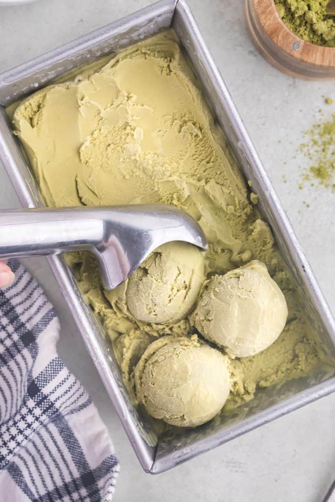 Overhead view of matcha ice cream in a metal loaf pan. Three scoops of ice cream have been pulled with the metal ice cream scooper and sit near the bottom of the container. There is a sprinkling of matcha powder to the right of the ice cream container and a blue and white linen to the left of the container.
