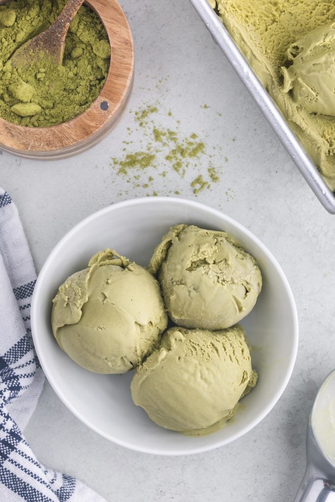 overhead shot of a small white bowl filled with three scoops of creamy green matcha ice cream. The upper left corner contains a wooden bowl of green matcha powder, some has spilled out over its edge. The top right corner is a metal loaf pan of matcha ice cream.