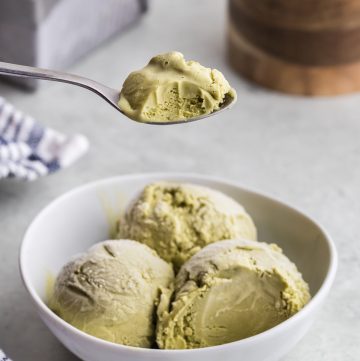 Side view of a small white bowl with three scoops of earthy green matcha ice cream in it. A spoon is held overhead with a spoonful of ice cream removed from one scoop and a bite taken out.
