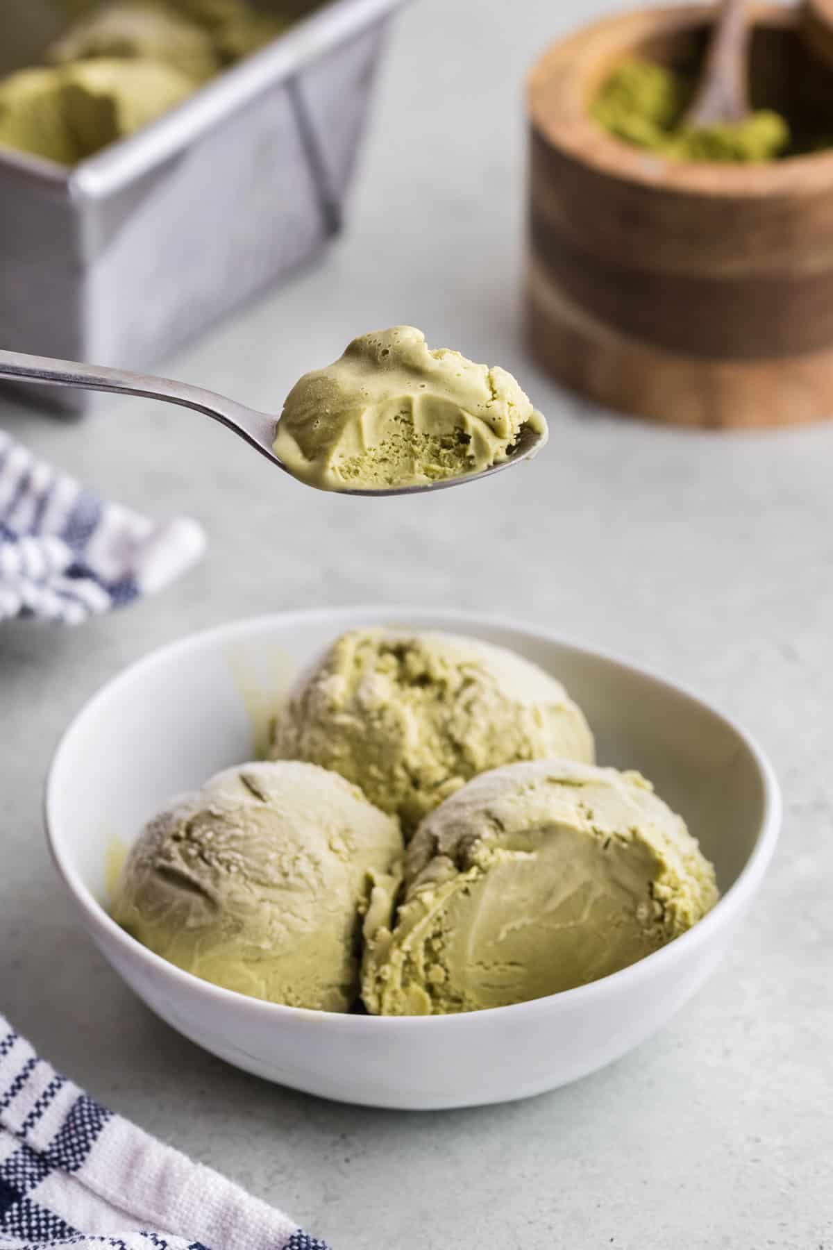 Side view of a small white bowl with three scoops of earthy green matcha ice cream in it. A spoon is held overhead with a spoonful of ice cream removed from one scoop and a bite taken out.