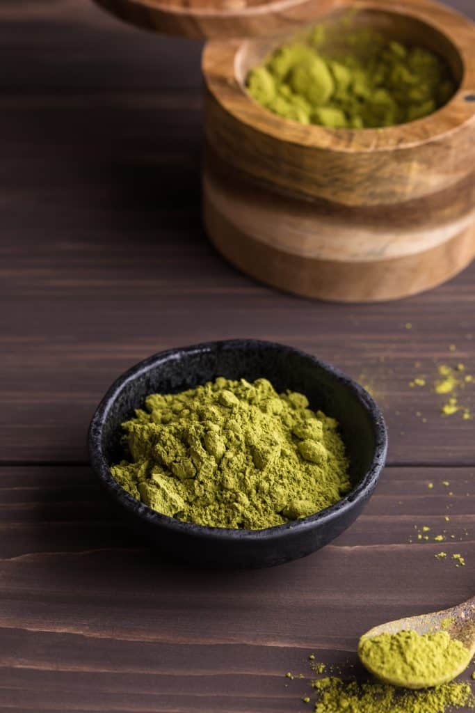 close up of bright green matcha powder in a small black earthenware dish. A larger wooden container is in the background with more matcha powder and a small pottery spoon filled with a scoop of matcha powder sits towards the bottom right of the earthenware bowl.