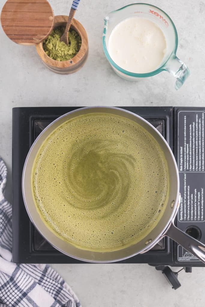 Overhead view of matcha, milk, eggs, and sugar woarming in a saucepan to form the custard base for matcha ice cream. The contents of the saucepan is an earthy green and it has frothy lighter green swirls in it.