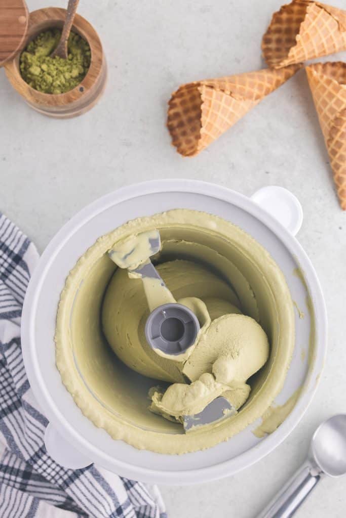 overhead view of the green matcha ice cream in the ice cream maker bowl just after it has finished churning. The ice cream is a light earthy green color and has the texture of soft serve ice cream. The top of the frame has a few waffle cones scattered around