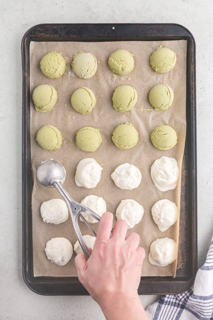 Overhead shot of a baking pan covered in a light brown sheet of parchment paper covered in neat rows of scooped ice cream balls with a hand holding an ice cream scooping dropping the last ice cream ball on.